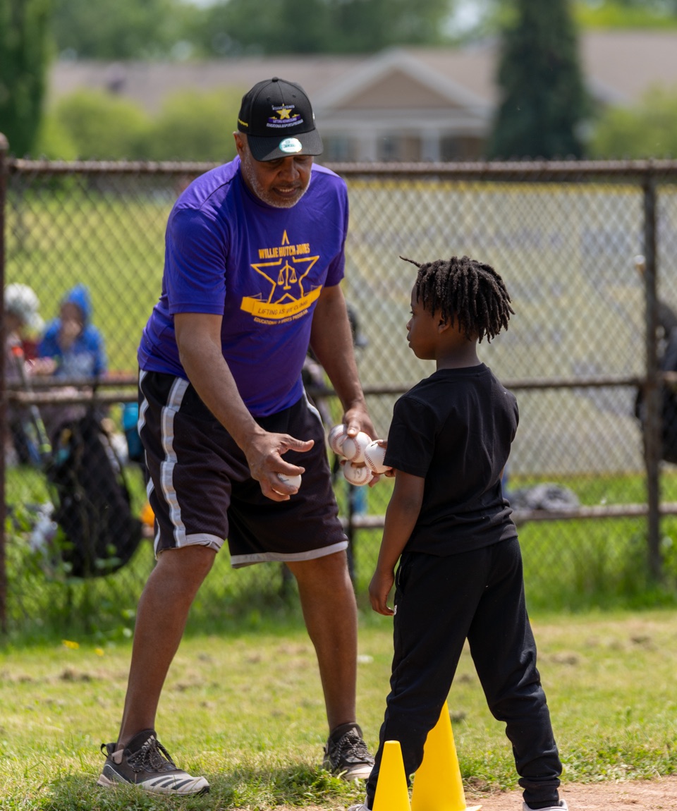 A WHJESP coach in a purple Willie Hutch Jones shirt hands baseballs to a young player during a youth baseball clinic.